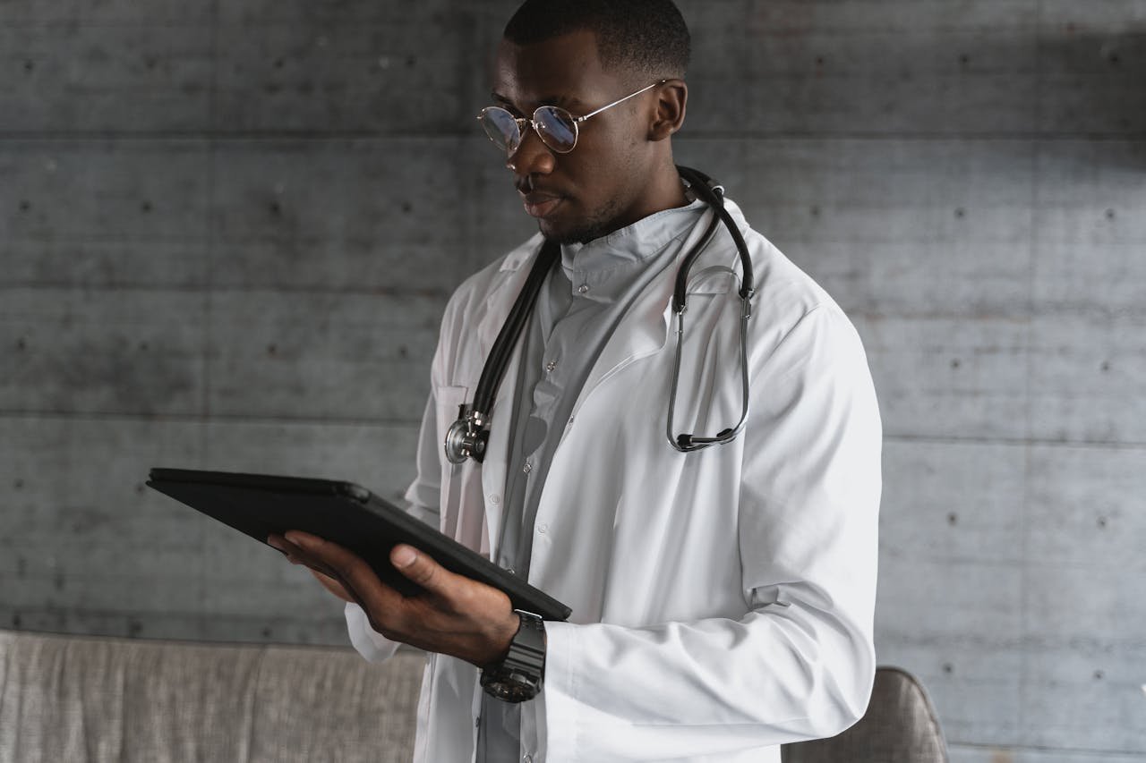 A healthcare professional using a tablet indoors, wearing a white coat and stethoscope.